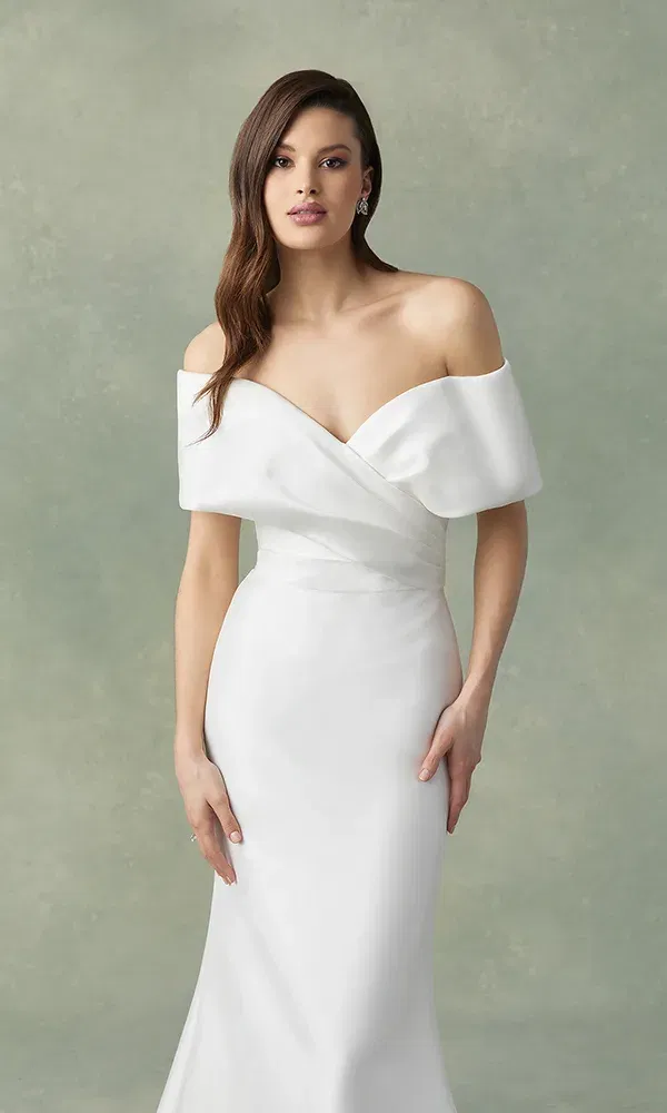 Ein schlichtes, aber edles Brautkleid mit schulterfreiem Ausschnitt umhüllt die schlanke Figur der Braut. Ihr Haar ist in sanfte Wellen gelegt, und sie trägt dezente Ohrringe, die den eleganten Look vervollständigen. Der neutrale Hintergrund lässt das makellose Weiß des Kleides besonders gut zur Geltung kommen.