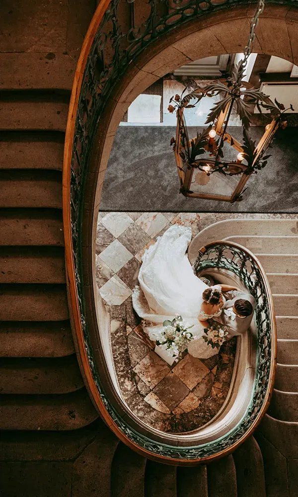 Das Brautpaar, von oben fotografiert, steht auf dem Boden einer prunkvollen Treppe. Der Brautschleier ist über die Treppenstufen ausgebreitet, während eine antike Laterne über ihnen hängt.
