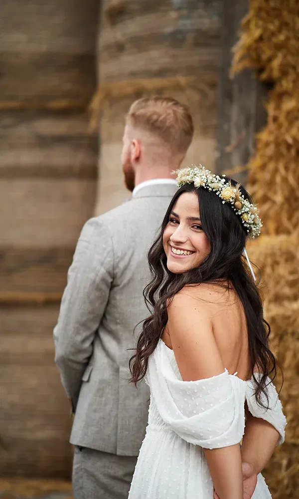 Eine Braut mit Blumenkranz dreht sich lächelnd zur Kamera um, während ihr Bräutigam mit dem Rücken zur Kamera steht. Der rustikale Heuhintergrund sorgt für eine ländliche Hochzeitsatmosphäre.