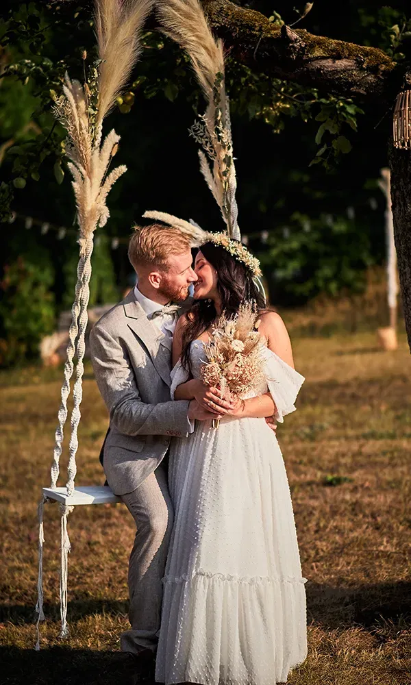Auf einer mit Pampasgras dekorierten Schaukel sitzt ein Hochzeitspaar eng beieinander. Die Braut hält einen Trockenblumenstrauß und trägt einen Blumenkranz, während der Bräutigam sie zärtlich ansieht.