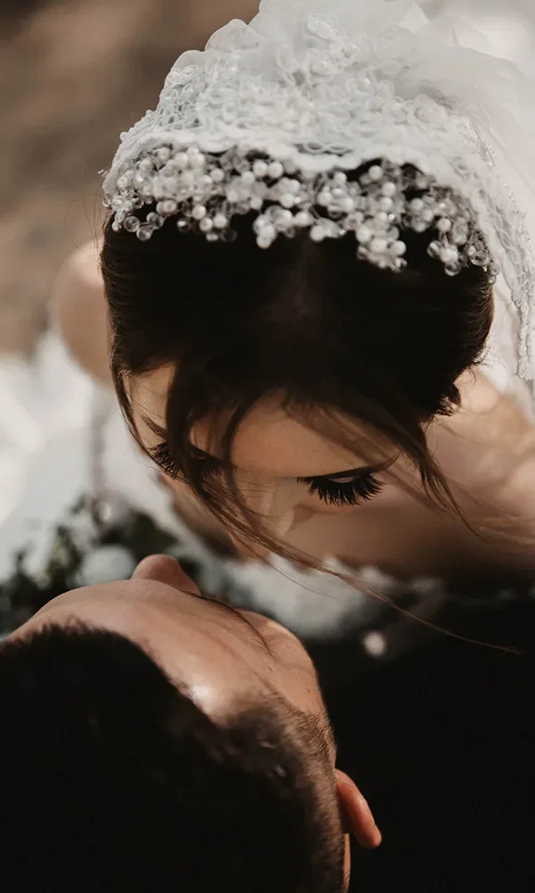 Die Braut mit einem verzierten, perlenbesetzten Schleier beugt sich in einer Nahaufnahme zu ihrem Bräutigam. Ihre langen Wimpern und der Schleier stehen im Fokus des Bildes. Der Bräutigam liegt entspannt und schaut sie an.