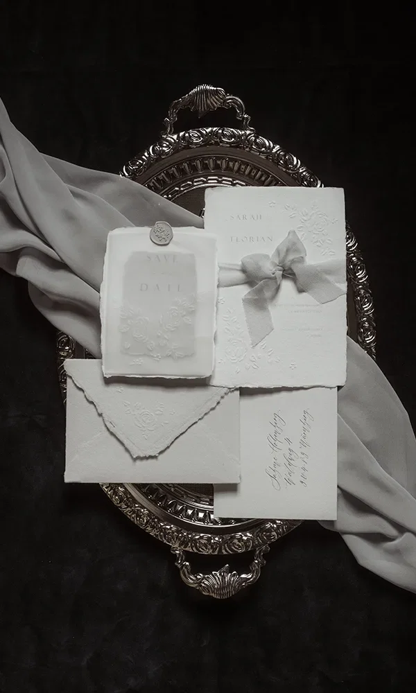Ein Set aus Hochzeitspapeterie, darunter Einladungen und Save-the-Date-Karten, liegt auf einem Silbertablett. Die Karten sind elegant arrangiert und von einem grauen Stoff dekorativ umgeben.