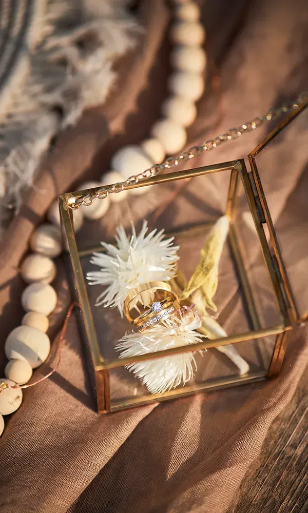 In einer stilvollen Glasbox liegen zwei goldene Eheringe auf weißen Trockenblumen. Die Szene ist auf einem natürlichen Stoff dekoriert, und eine Holzkette verleiht der Anordnung einen rustikalen Look.