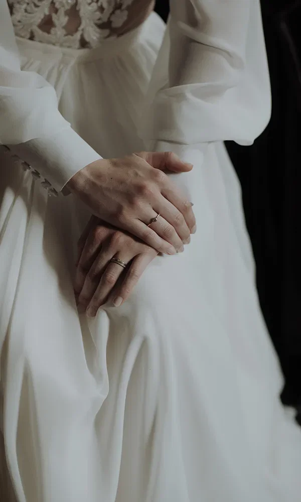 Hände der Braut in einer ruhigen Pose, die auf ihrem weißen Brautkleid liegen. Der goldene Ehering an ihrem Finger fällt ins Auge, während das fließende Material des Kleides den Hintergrund bildet.