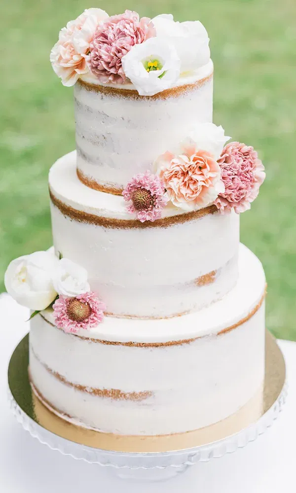 Eine elegante Hochzeitstorte im rustikalen Stil mit dünner Buttercreme-Beschichtung und frischen Blumen in Rosa und Weiß ziert eine dreistöckige Semi-Naked Torte auf einem gläsernen Tortenständer.