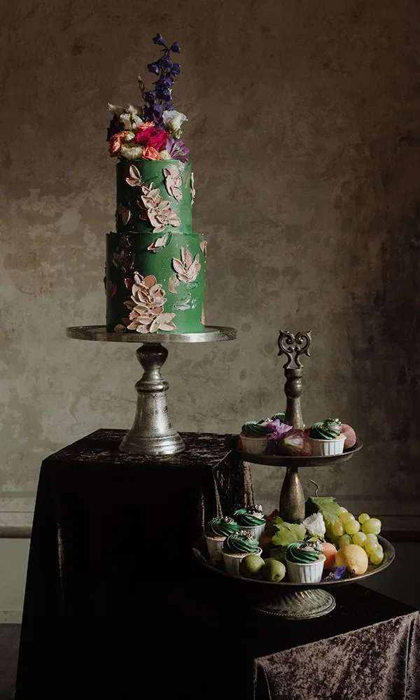 Eine dreistöckige grüne Torte mit kunstvollen Blumenmustern steht im Mittelpunkt. Auf einem zweiten Tisch sind Cupcakes und frisches Obst wie Trauben und Zitrusfrüchte auf einer Etagere angerichtet.