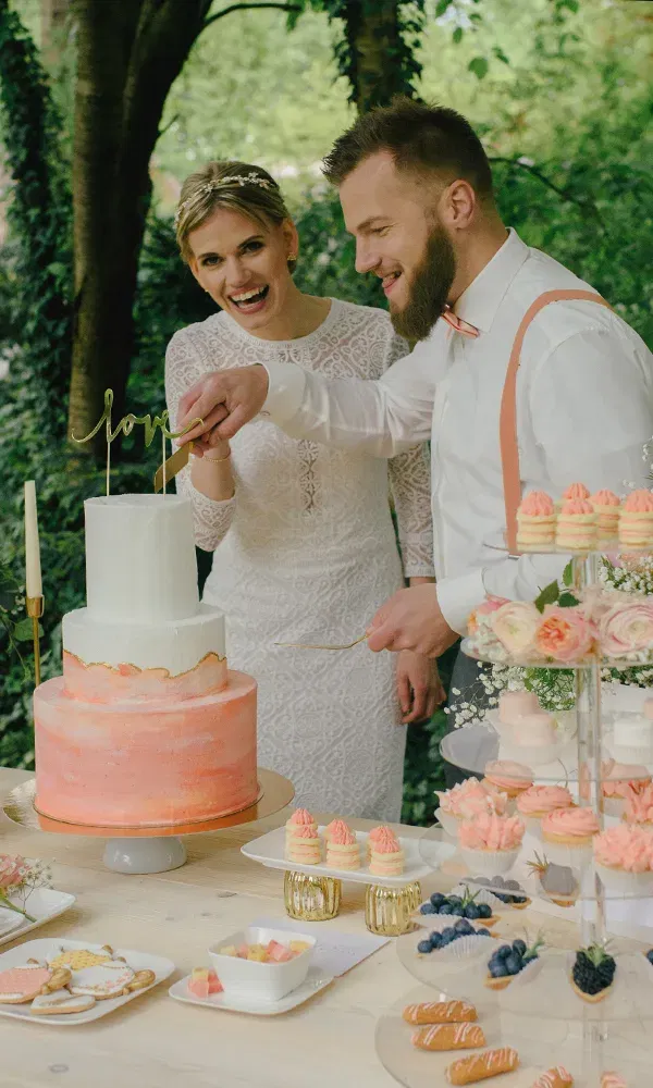 Ein glückliches Brautpaar, das eine elegante zweistöckige Hochzeitstorte anschneidet. Die Torte ist in einem zarten Rosa gehalten, und der Tisch ist mit süßen Köstlichkeiten und Blumen dekoriert.
