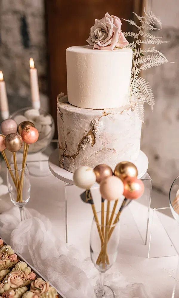 Die zweistöckige Hochzeitstorte, verziert mit Rosen und filigranen Details, steht neben rosé- und goldfarbenen Cake-Pops auf einem Tisch. Der Tisch ist mit weißem Stoff dekoriert und von Kerzen erleuchtet.