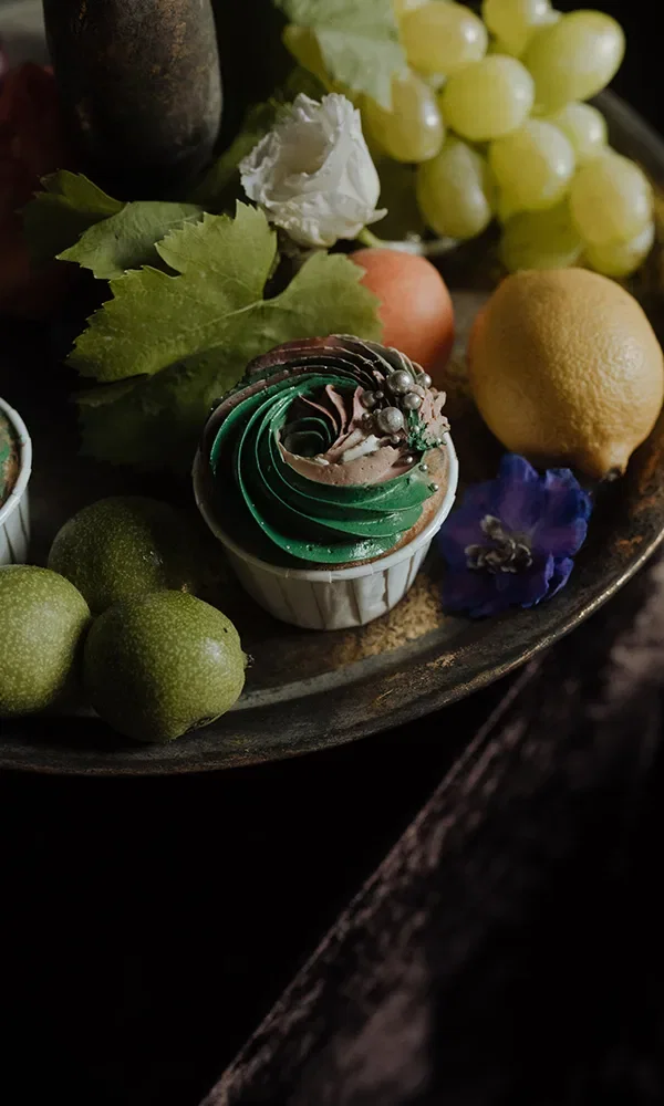 Ein Cupcake mit kunstvollem, grünem Zuckerguss ist zusammen mit Früchten wie Trauben, Limetten und einer Zitrone auf einem Tablett arrangiert. Blätter und Blumen ergänzen die elegante Präsentation.
