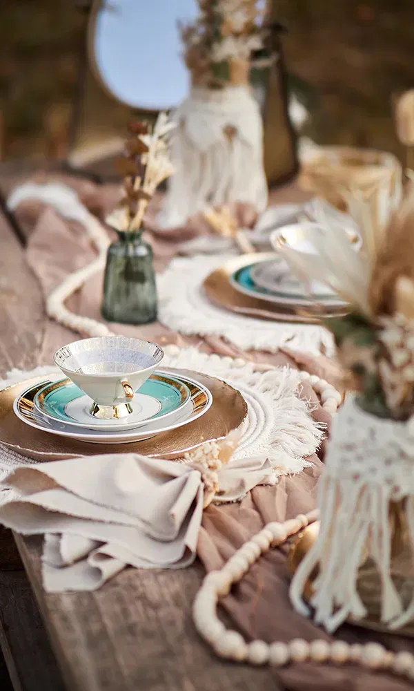 Ein dekorativer Esstisch zeigt eine stilvolle Anordnung von Geschirr, inklusive einer Teetasse auf einer goldenen Unterlage. Umgeben von Trockenblumen und natürlichen Stoffen, ist der Tisch für eine Hochzeit im Boho-Stil geschmückt.