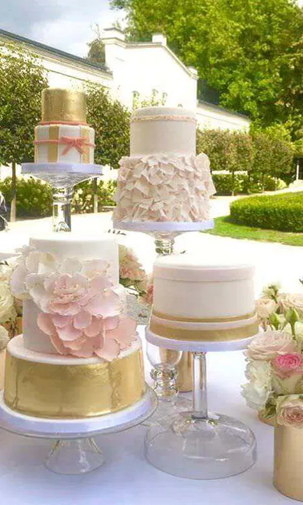 Verschiedene Hochzeitstorten, die mit goldenen und weißen Dekorationen verziert sind, stehen auf einem Tisch. Die Torten zeigen unterschiedliche Designs, einige mit Blumen und andere mit schlichten, eleganten Mustern.