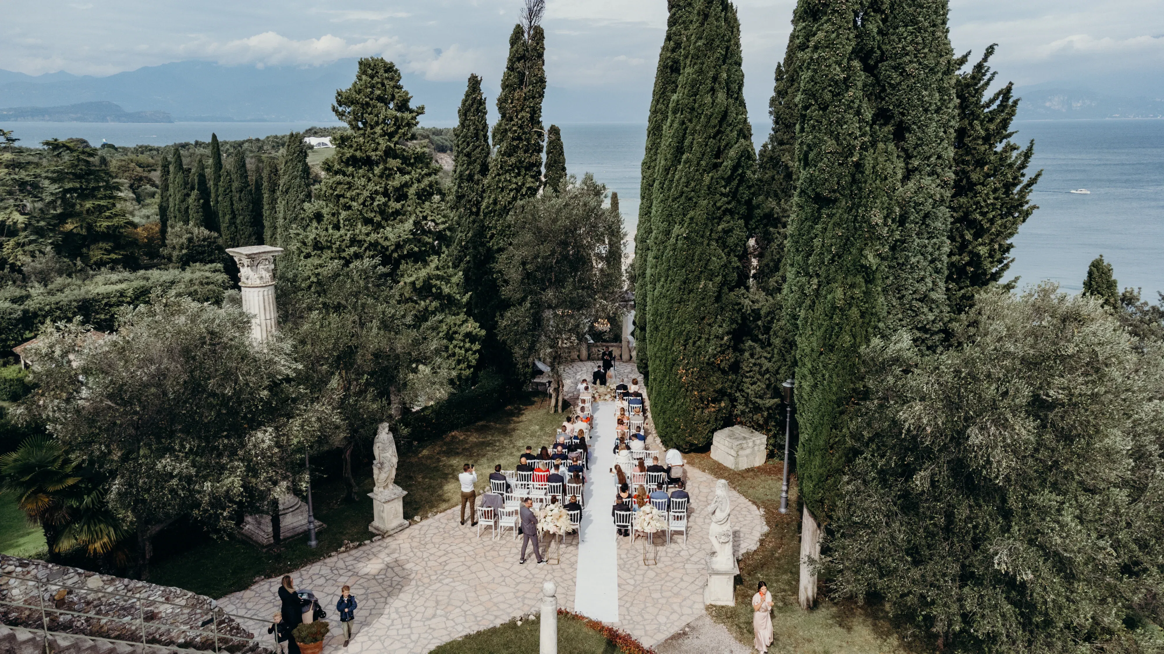Eine idyllische Outdoor-Hochzeit mit Meerblick, umgeben von alten Bäumen und Skulpturen, die Gäste sitzen auf weißen Stühlen entlang eines Steinwegs.