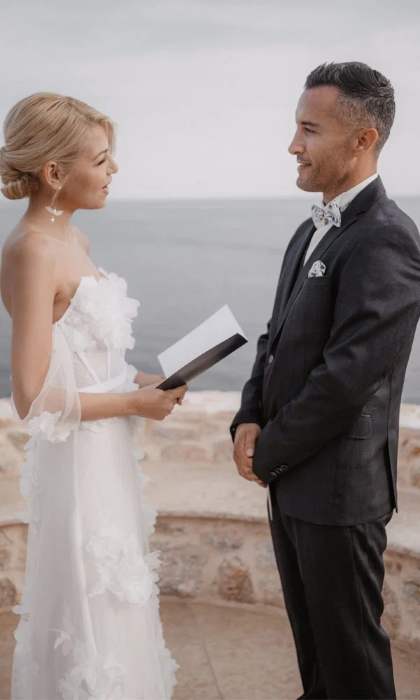 Eine romantische Szene bei einer Strandhochzeit. Die Braut, gekleidet in einem weißen Kleid, liest aus einem Notizbuch ihre Gelübde vor, während der Bräutigam in einem eleganten Anzug lächelnd zuhört. Im Hintergrund ist das Meer zu sehen.