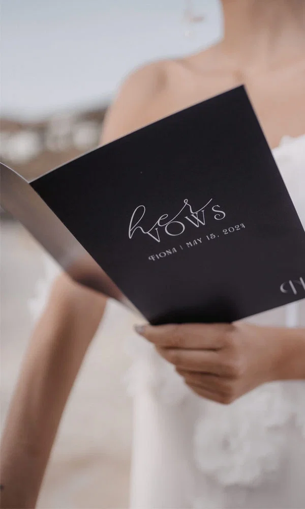 Die Braut liest ihre Gelübde aus einem schwarzen Heft mit der Aufschrift 'Her Vows' auf der Titelseite. Der elegante, schlichte Stil des Hefts wird in den Mittelpunkt gerückt.