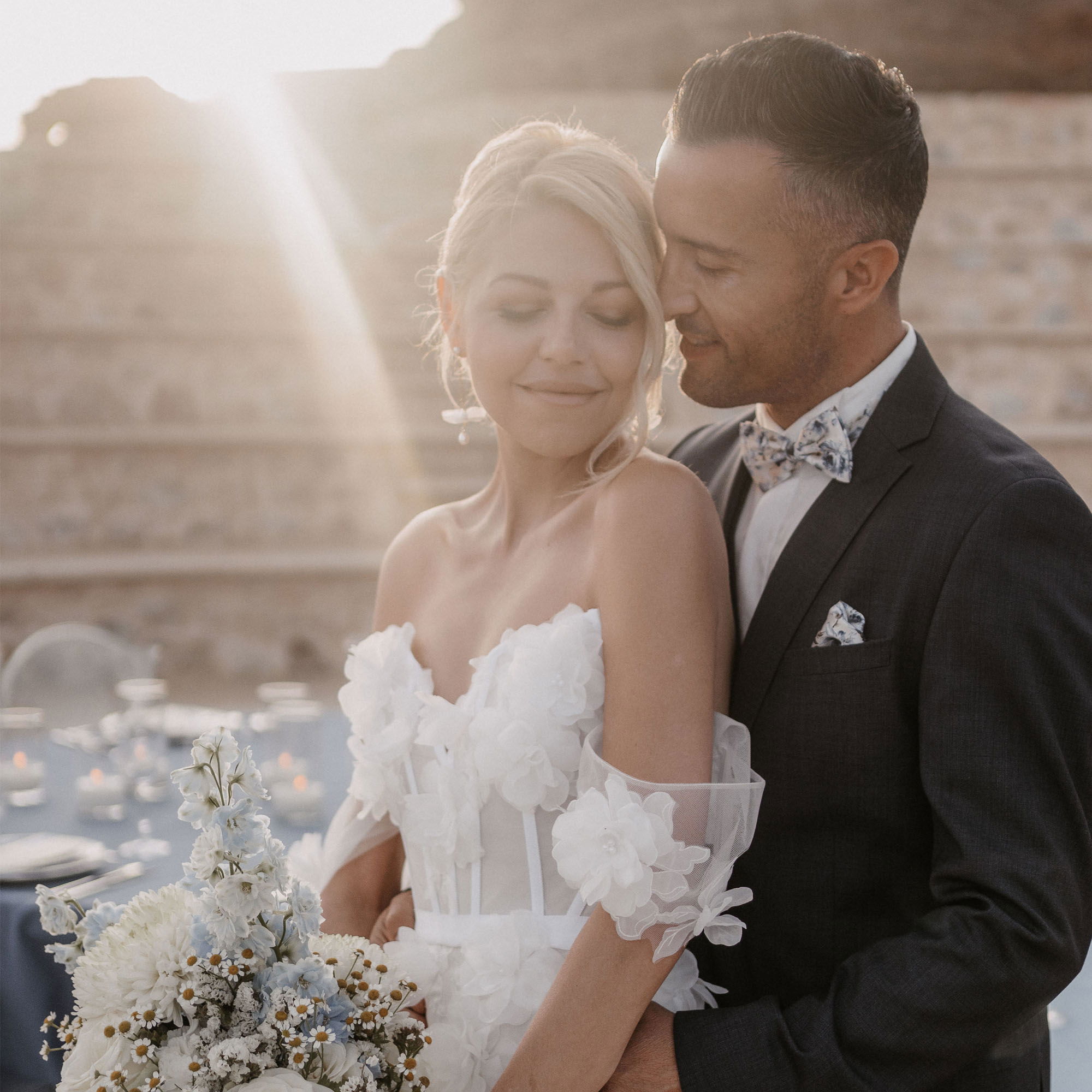 Die Braut und der Bräutigam posieren zärtlich im warmen Licht der untergehenden Sonne. Die Braut hat ein detailliertes weißes Kleid an und hält einen Strauß aus weißen und blauen Blumen. Der Bräutigam hält sie fest umschlungen und lächelt, während er seine Stirn fast an ihre legt. Die Steintreppe im Hintergrund verstärkt die romantische Atmosphäre.