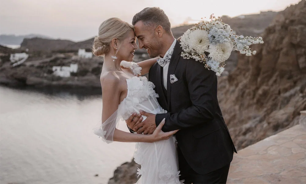 Ein glückliches Brautpaar posiert am Rande einer felsigen Küste. Die Braut hält einen üppigen Strauß aus weißen Blumen und trägt ein elegantes Kleid, während der Bräutigam einen dunklen Anzug mit Fliege trägt.