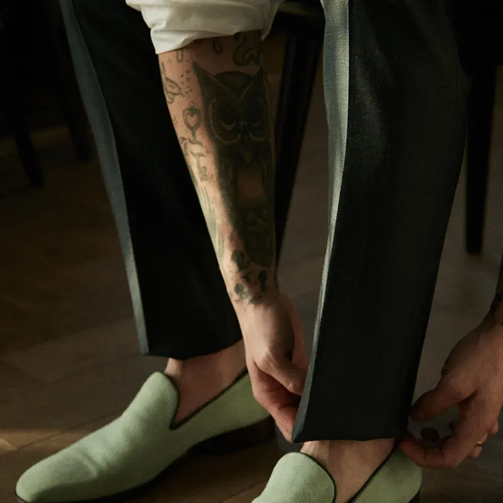 Ein tätowierter Bräutigam, gekleidet in dunkle Hosen, beugt sich nach vorne, um seine grünen Slipper anzuziehen. Sein Eulentattoo ist gut sichtbar, und das Sonnenlicht beleuchtet den Holzboden sanft.