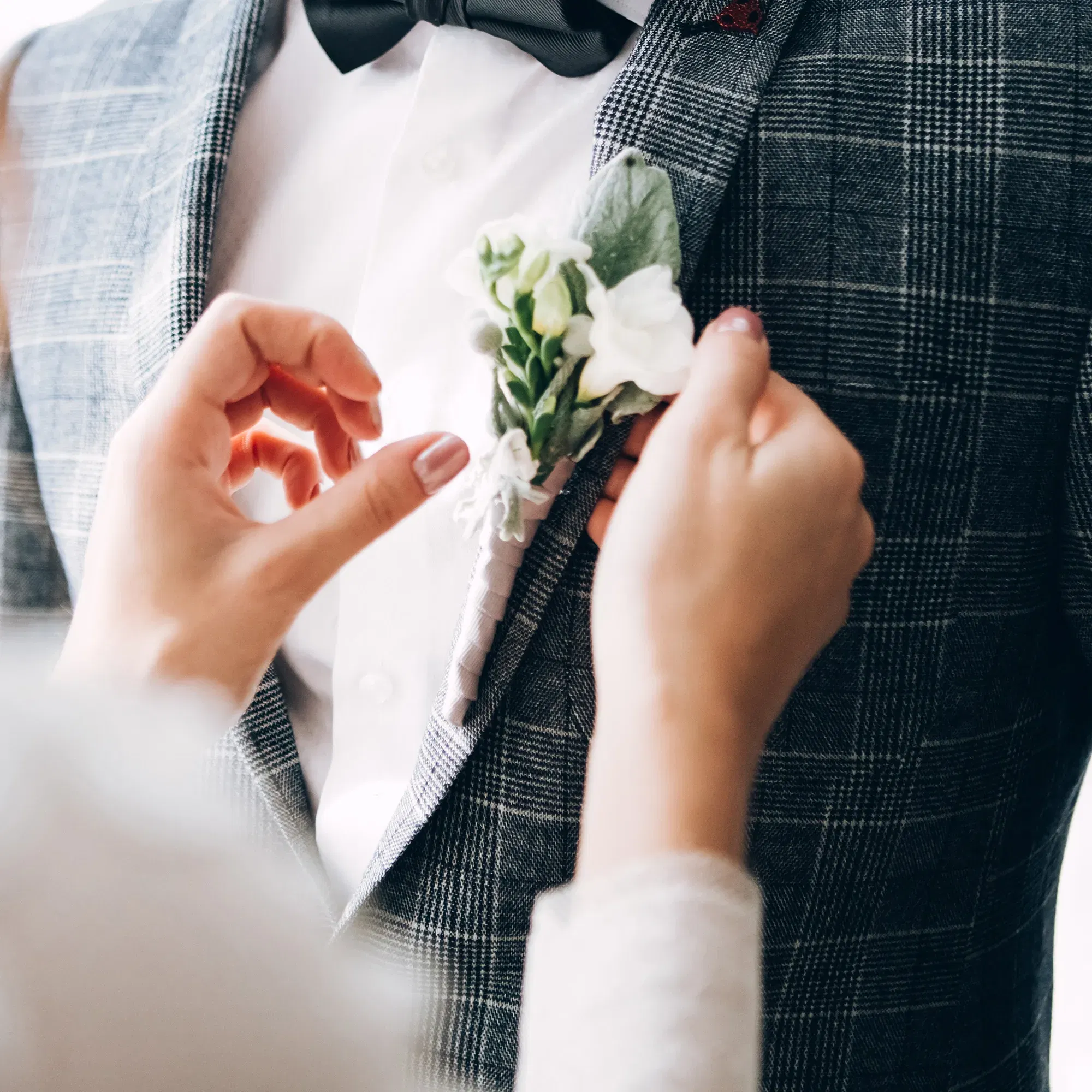 Eine Boutonnière wird am Revers eines Bräutigams mit Karoanzug befestigt. Die weiße Blume und die schwarze Fliege des Bräutigams sorgen für einen klassischen, stilvollen Hochzeitslook.