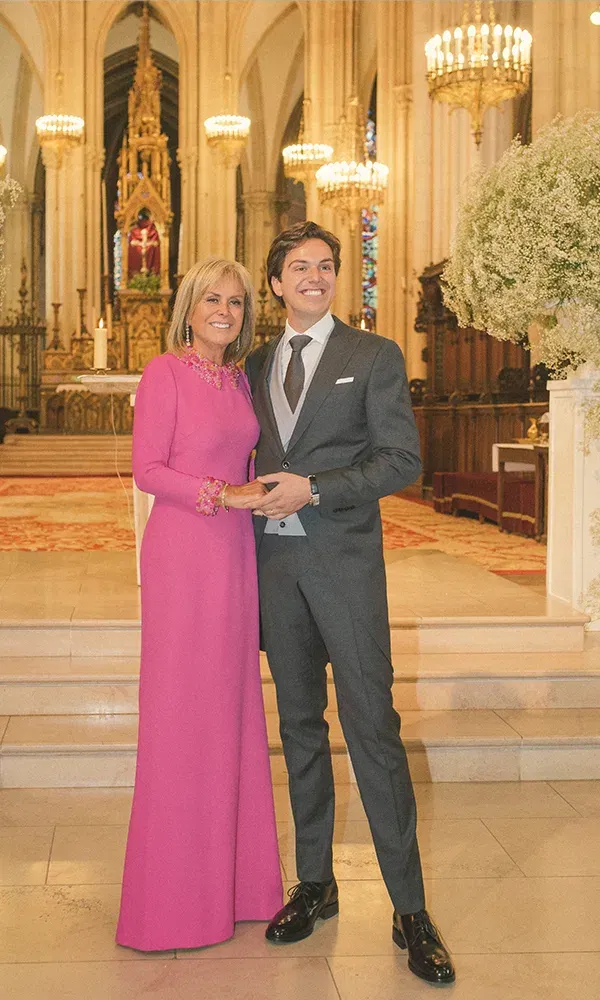 Der Bräutigam und seine Mutter lächeln fröhlich in einer großen Kirche. Die Mutter trägt ein auffälliges, pinkes Kleid, und der Hintergrund zeigt den goldverzierten Altar und beeindruckende Kronleuchter.