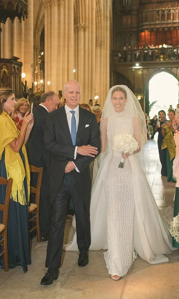 Eine strahlende Braut wird von ihrem Vater in einer beeindruckenden Kirche den Gang entlanggeführt. Die Gäste beobachten den Einzug, und die prächtigen Säulen und Kronleuchter im Hintergrund verstärken die festliche Atmosphäre.