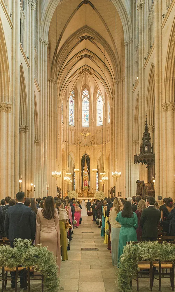 Eine Hochzeit findet in einer prachtvollen Kathedrale statt, die mit hohen Bögen und bunten Glasfenstern dekoriert ist. Die Gäste sitzen in den Bänken, und am Altar steht das Brautpaar, während die Zeremonie stattfindet.