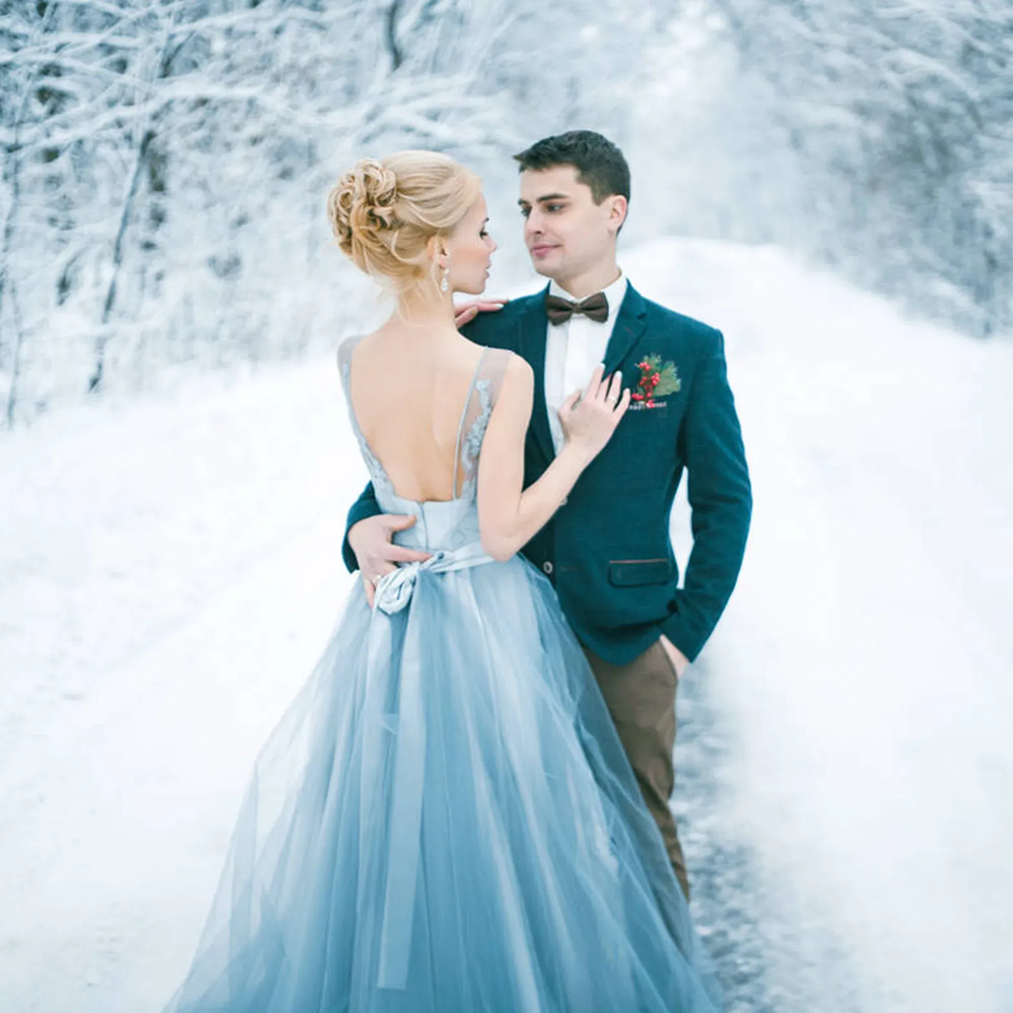 Ein Paar posiert romantisch in einer verschneiten Landschaft. Die Frau trägt ein langes, blaues Tüllkleid mit tiefem Rückenausschnitt, während der Mann einen eleganten Blazer und eine Fliege trägt. Der Weg ist von schneebedeckten Bäumen gesäumt.