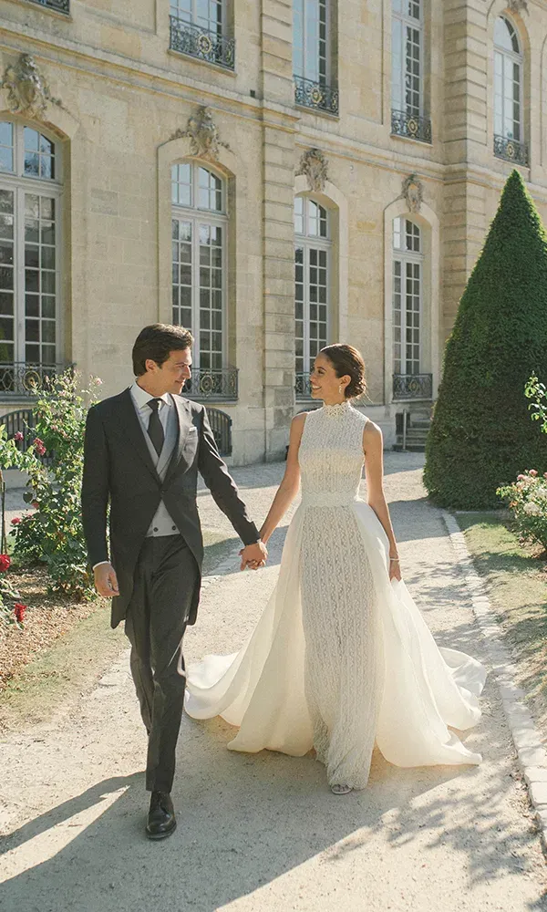 Ein Hochzeitspaar geht Hand in Hand vor einem historischen Schloss entlang. Die Braut trägt ein elegantes, hochgeschlossenes Brautkleid mit einem langen Umhang, und der Bräutigam ist in einem stilvollen Anzug gekleidet.