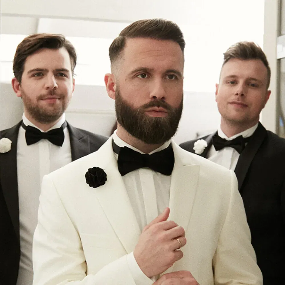 Der Bräutigam in einem stilvollen weißen Smoking steht im Vordergrund und richtet seine Kleidung. Hinter ihm stehen zwei Groomsmen in schwarzen Anzügen, die gemeinsam eine ernste, aber elegante Atmosphäre verbreiten.