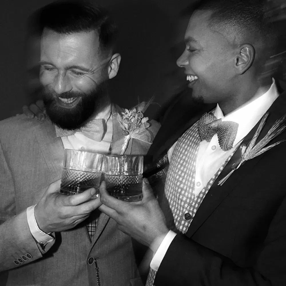 Der Bräutigam und sein Trauzeuge lachen herzlich und stoßen ihre Whiskeygläser an. Diese schwarz-weiß Aufnahme zeigt die beiden Männer in ihren eleganten Anzügen, die Freude und Verbundenheit während der Hochzeitsfeier teilen.