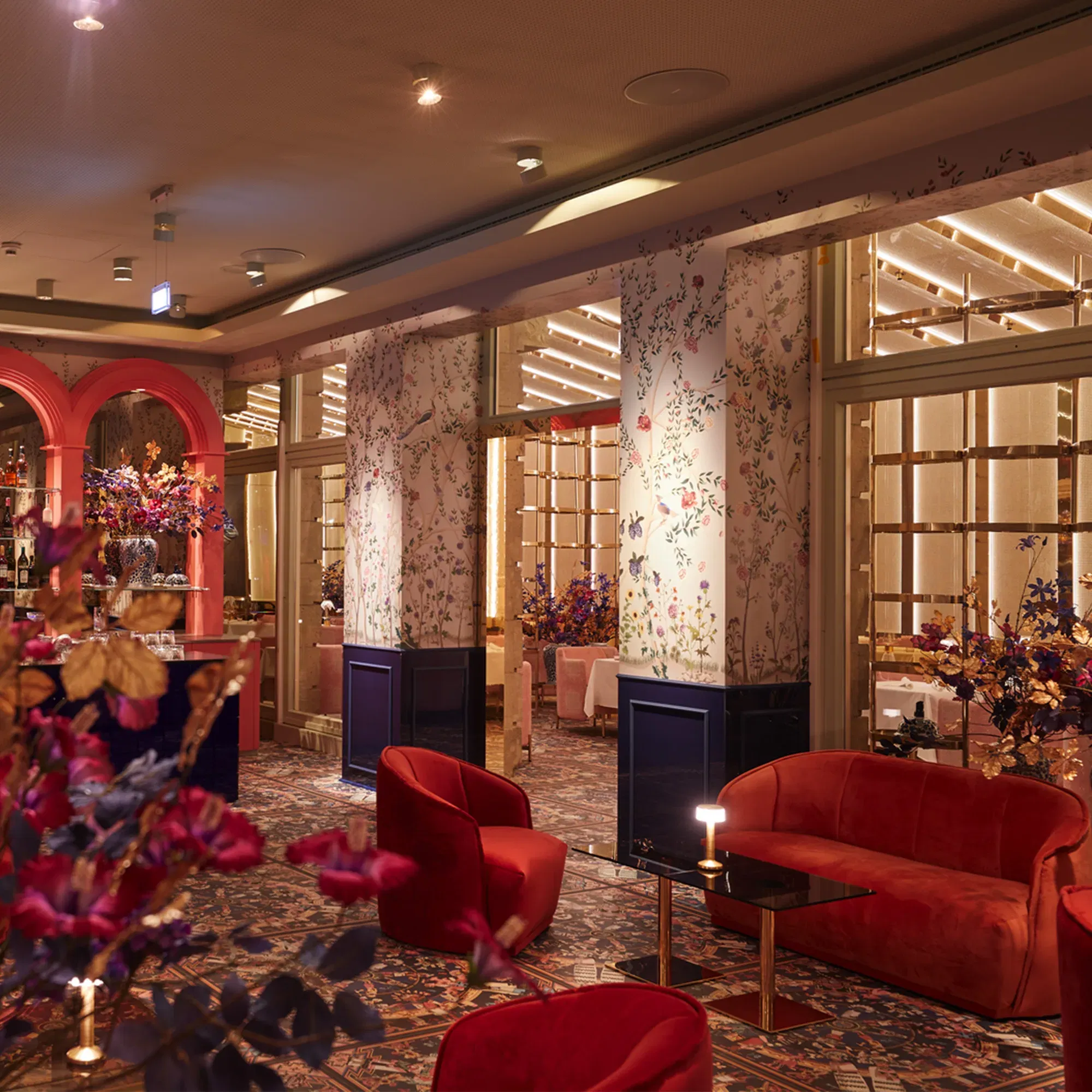 Eine schicke Lounge mit roten Samtsofas und modernen Blumenarrangements. Die Wände sind mit floralen Mustern dekoriert, und die Beleuchtung schafft eine gemütliche, luxuriöse Atmosphäre.