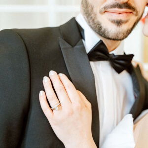 Die Hand der Braut ruht zärtlich auf der Schulter des Bräutigams in seinem schwarzen Hochzeitsanzug. Ihr Diamantring wird durch das weiche Licht betont, während der Bräutigam dezent im Hintergrund bleibt.