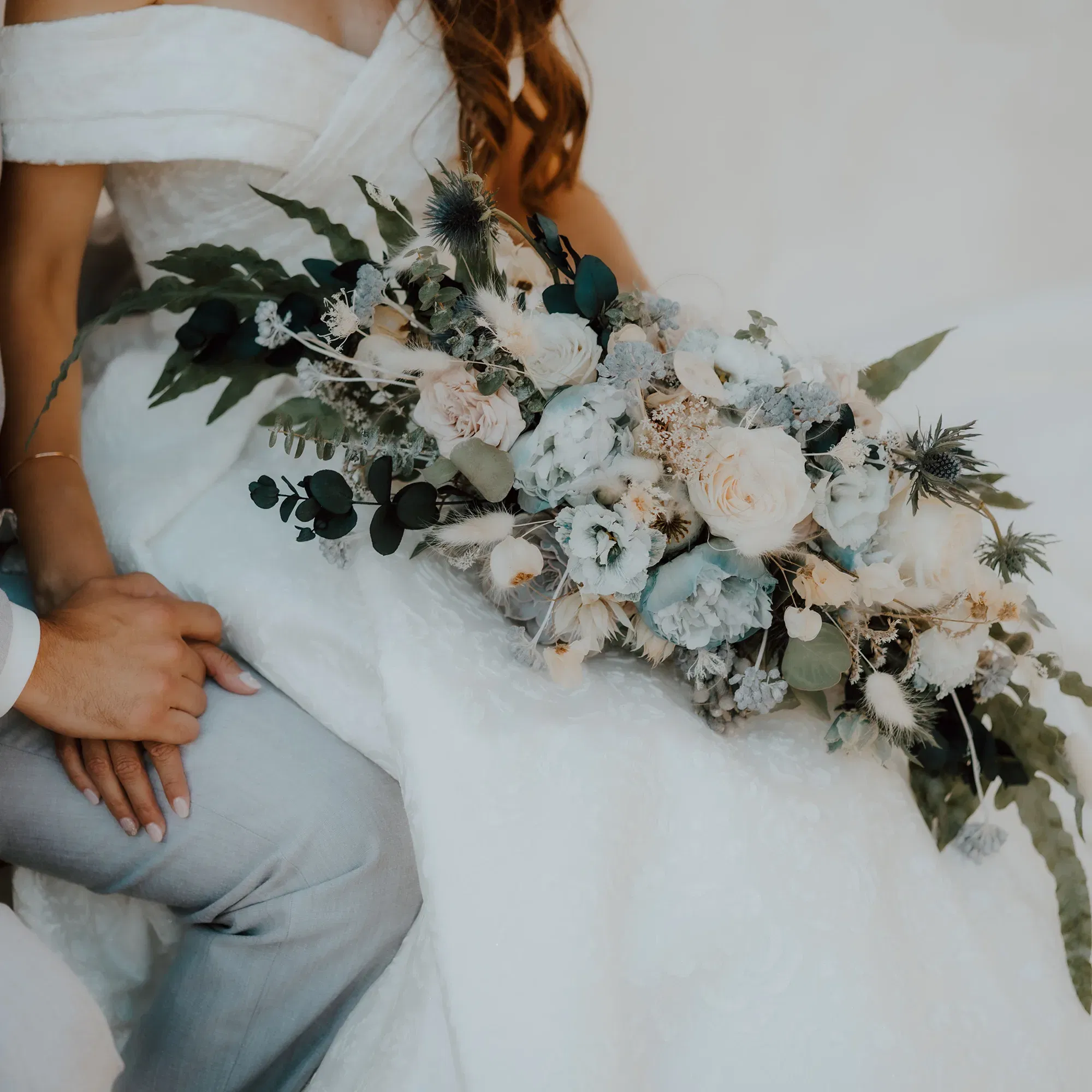 Der elegante Brautstrauß, bestehend aus weißen Rosen und grünen Eukalyptusblättern, ruht auf dem Schoß der Braut in einem aufwendigen weißen Hochzeitskleid. Ihr Partner hält liebevoll ihre Hand.
