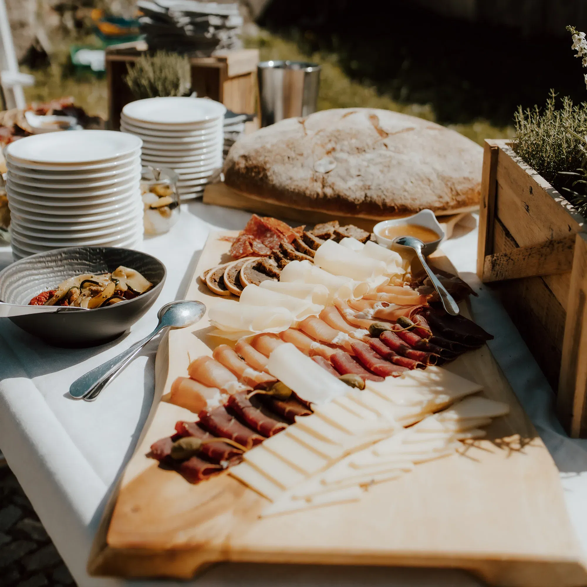 Eine elegante Käse- und Wurstplatte, kunstvoll angerichtet auf einem Holzbrett, wird auf einem Buffet präsentiert, zusammen mit frischem Brot und Beilagen in Schüsseln.