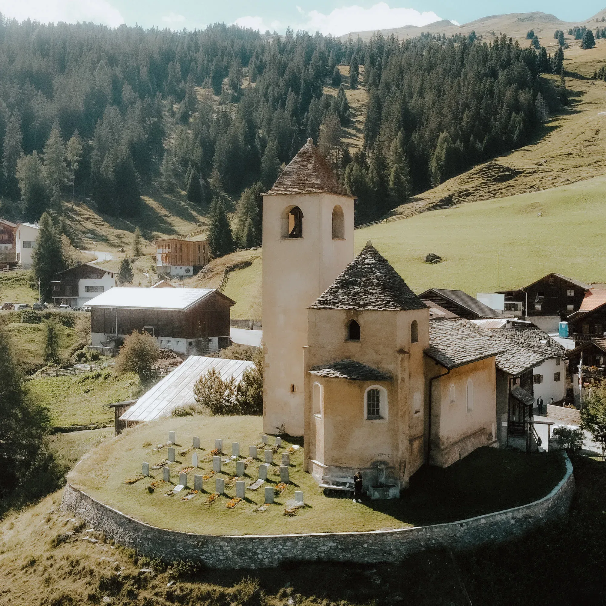 Eine kleine Kirche mit einem Friedhof liegt idyllisch inmitten einer hügeligen Landschaft, umgeben von Bauernhäusern und einem dichten Wald am Horizont.
