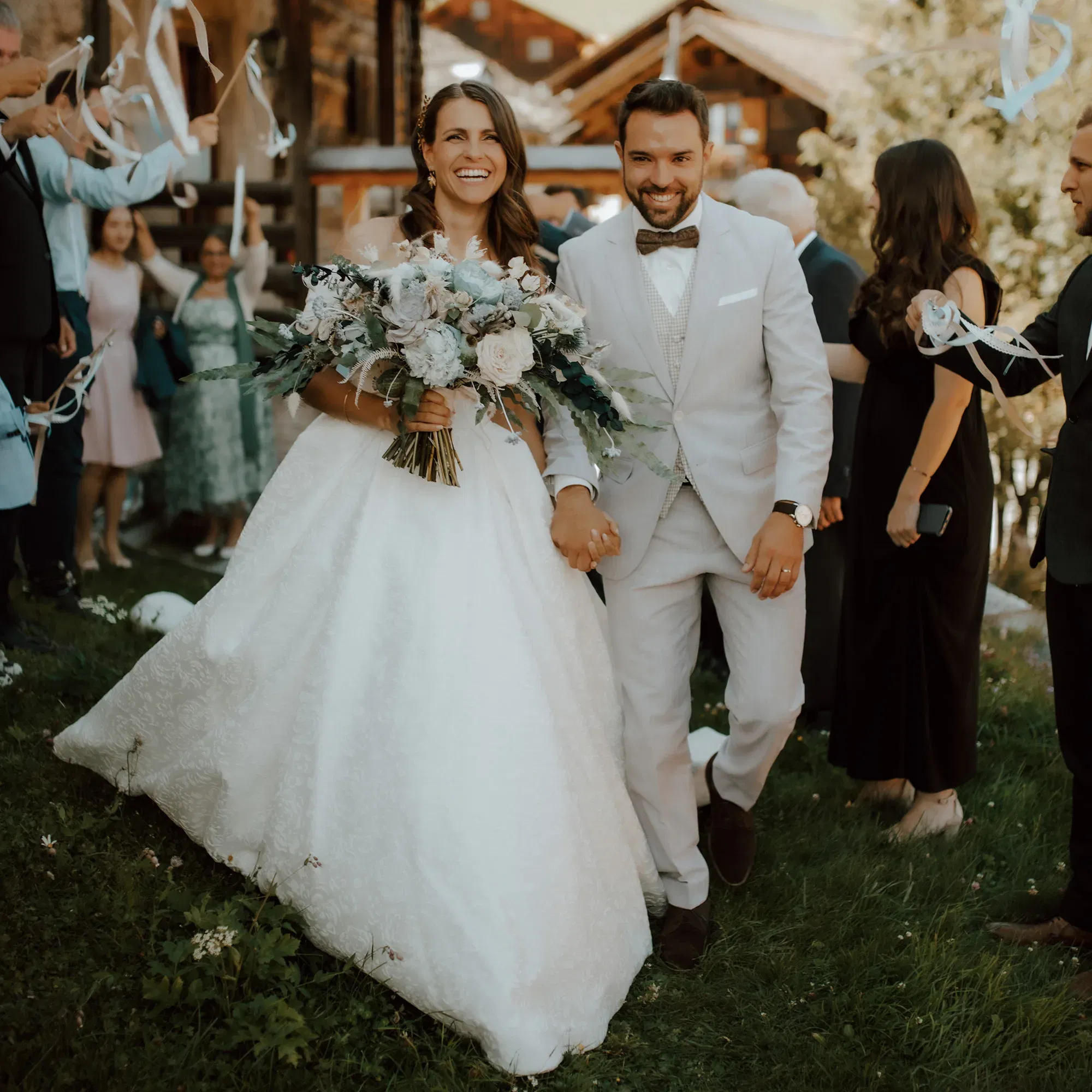 Ein glückliches Brautpaar läuft Hand in Hand durch die jubelnde Menge, während die Hochzeitsgäste weiße Bänder schwenken. Die Braut trägt ein prachtvolles weißes Kleid und einen großen Strauß pastellfarbener Blumen.