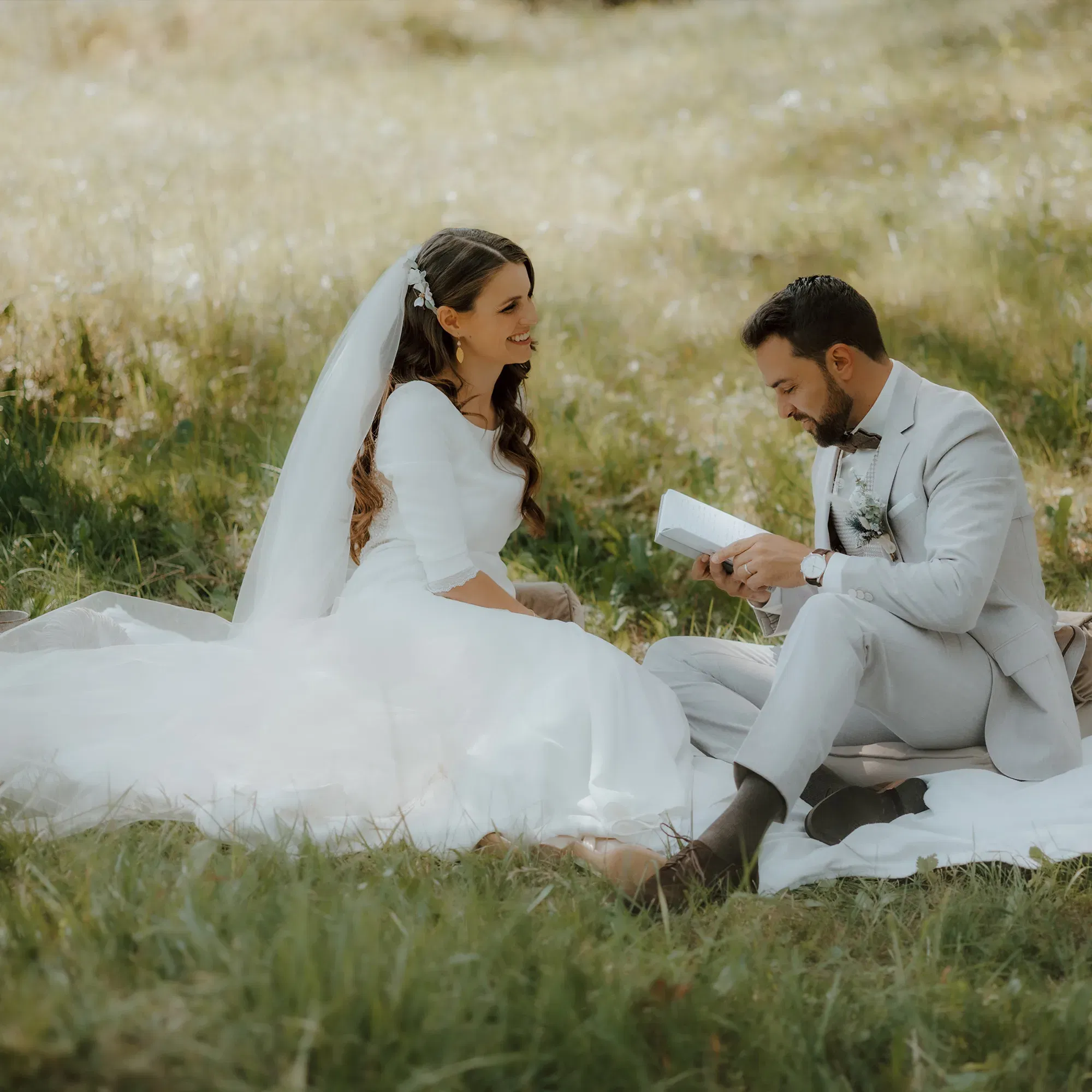 Das Brautpaar sitzt auf einer Picknickdecke in einer blühenden Wiese. Der Bräutigam liest der Braut, die ein langes weißes Kleid und einen Schleier trägt, liebevoll aus einem Brief vor.