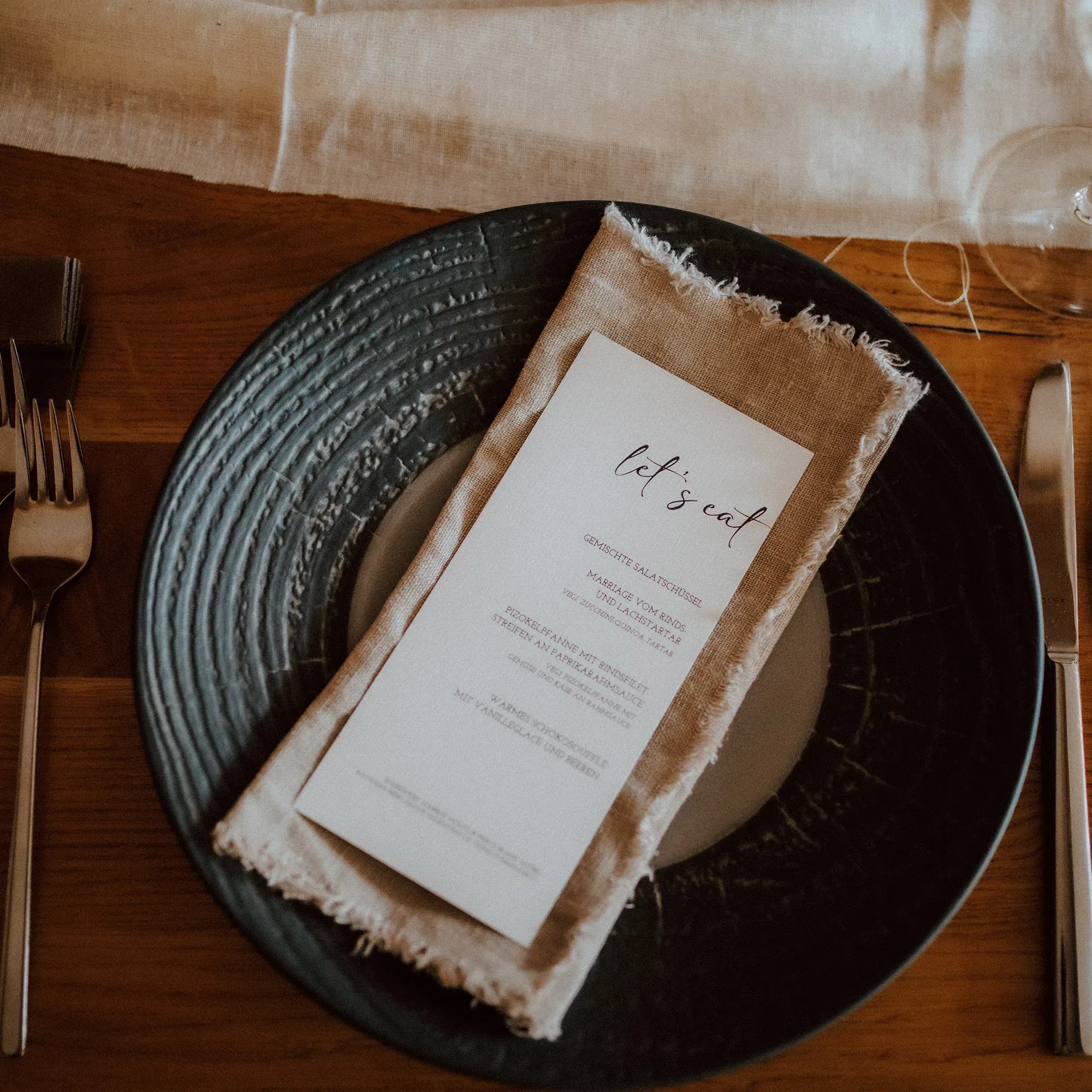 Eine elegante Tischdekoration mit einem schwarzen Teller, einer bestickten Serviette und einer Menükarte mit handgeschriebener Schrift, die das Hochzeitsmenü präsentiert.