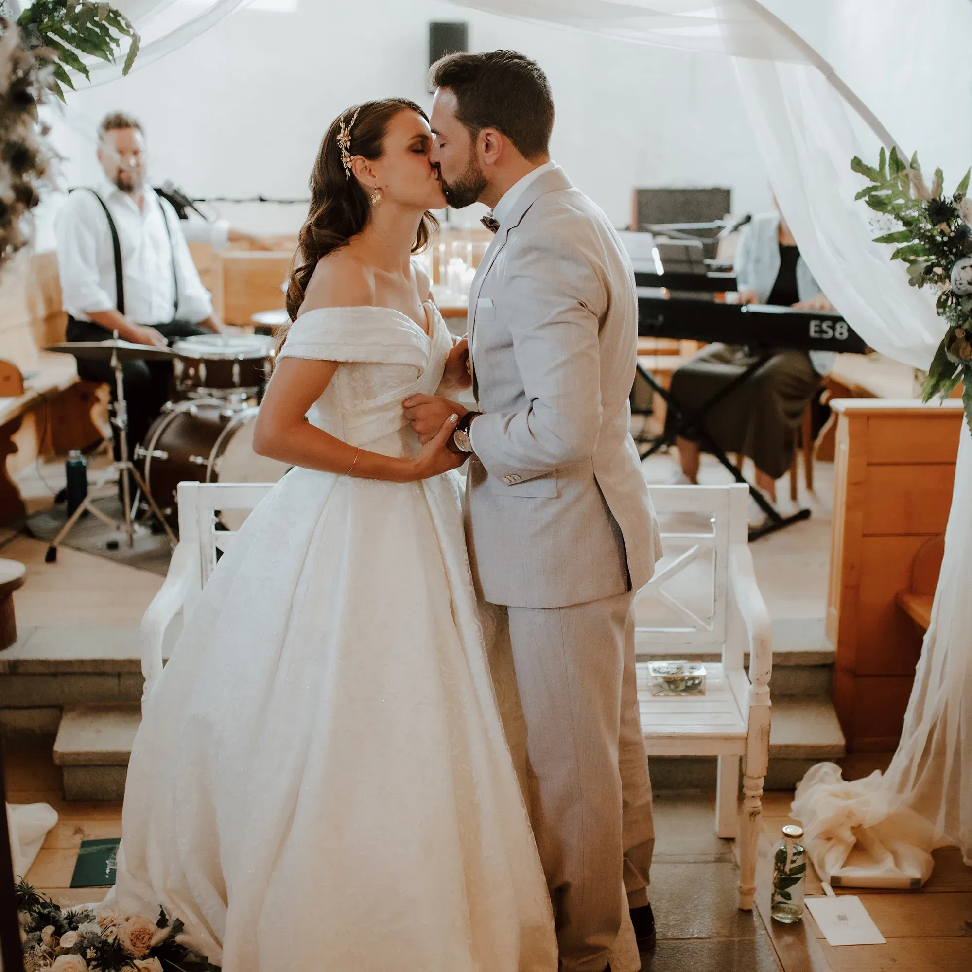 Während der Hochzeitszeremonie küsst sich das Brautpaar innig, umgeben von romantischer Atmosphäre. Die Braut trägt ein voluminöses weißes Hochzeitskleid, der Bräutigam einen grauen Anzug. Eine Band spielt dezente Musik im Hintergrund.
