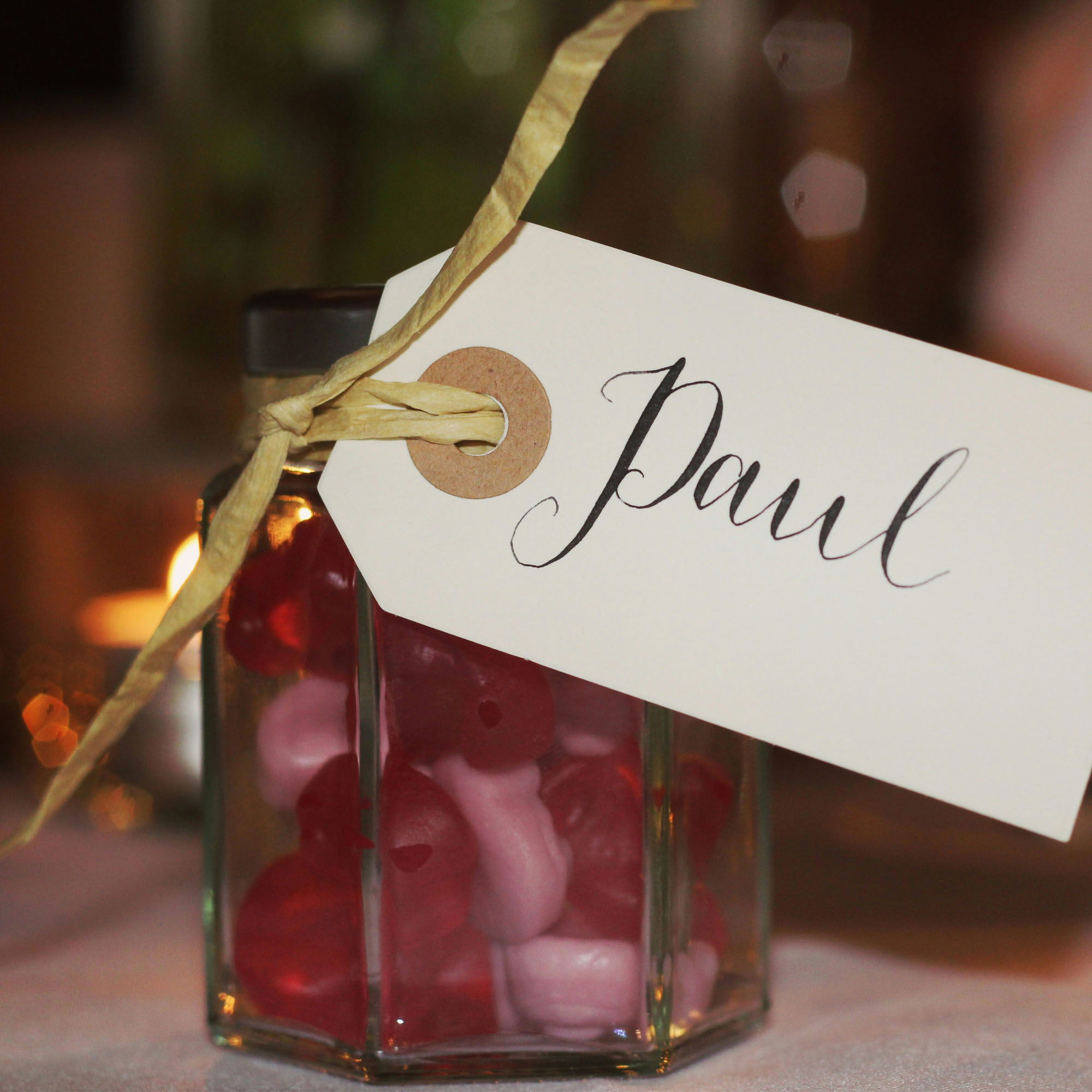 Ein rechteckiges Glas mit Süßigkeiten, hauptsächlich in den Farben Rot und Rosa, ist mit einem beige-farbenen Band und einem Etikett versehen, auf dem in geschwungener Handschrift "Paul" geschrieben steht. Der unscharfe Hintergrund deutet eine stimmungsvolle Beleuchtung an.