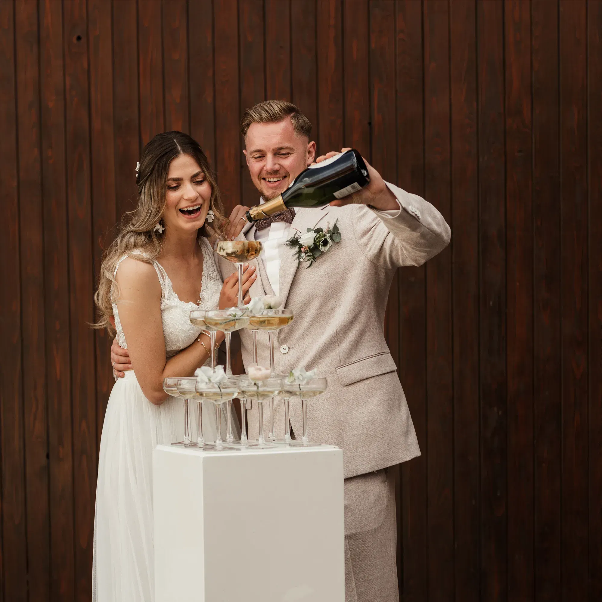Das Brautpaar gießt Champagner in eine Pyramide von Gläsern. Beide wirken glücklich und lachen, während sie den Moment feiern. Die Braut trägt ein weißes Kleid, der Bräutigam einen hellen Anzug.