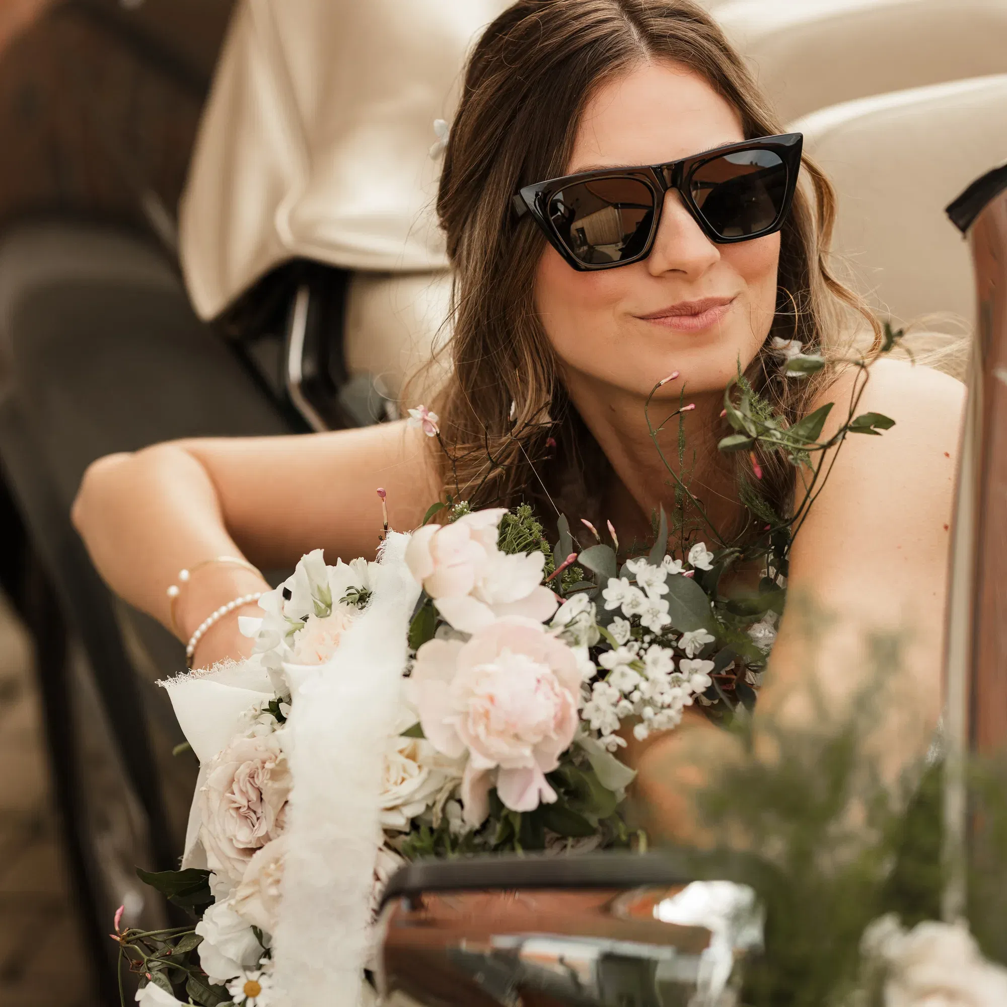 Eine lächelnde Braut mit großen Sonnenbrillen sitzt an einem Auto und hält einen wunderschönen Blumenstrauß aus hellen Blumen. Ihre Haare fallen locker über ihre Schultern.