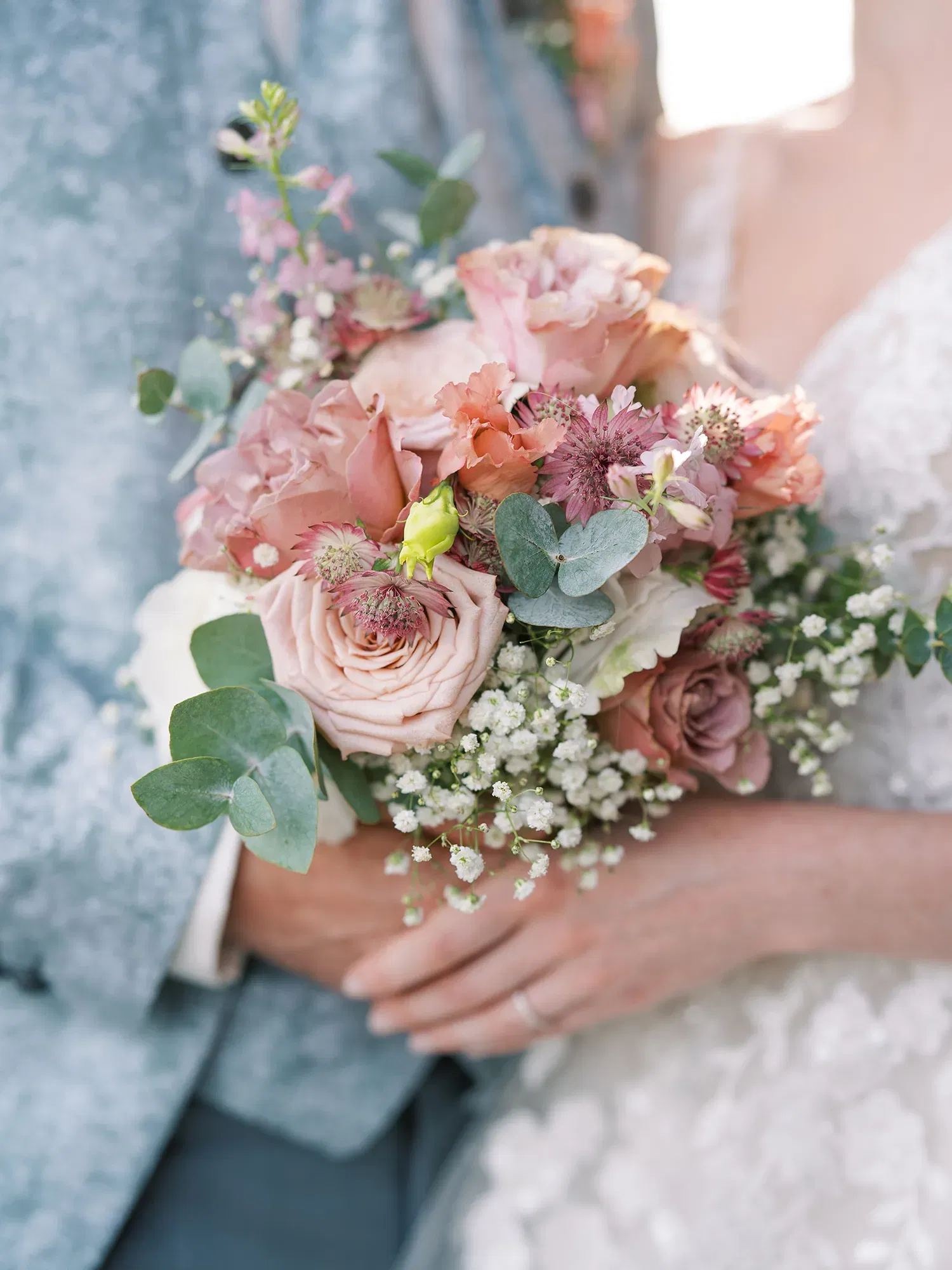 Detailaufnahme eines romantischen Brautstraußes aus Rosen und Eukalyptus, der von einer Braut und einem Bräutigam in eleganter Hochzeitskleidung gehalten wird.