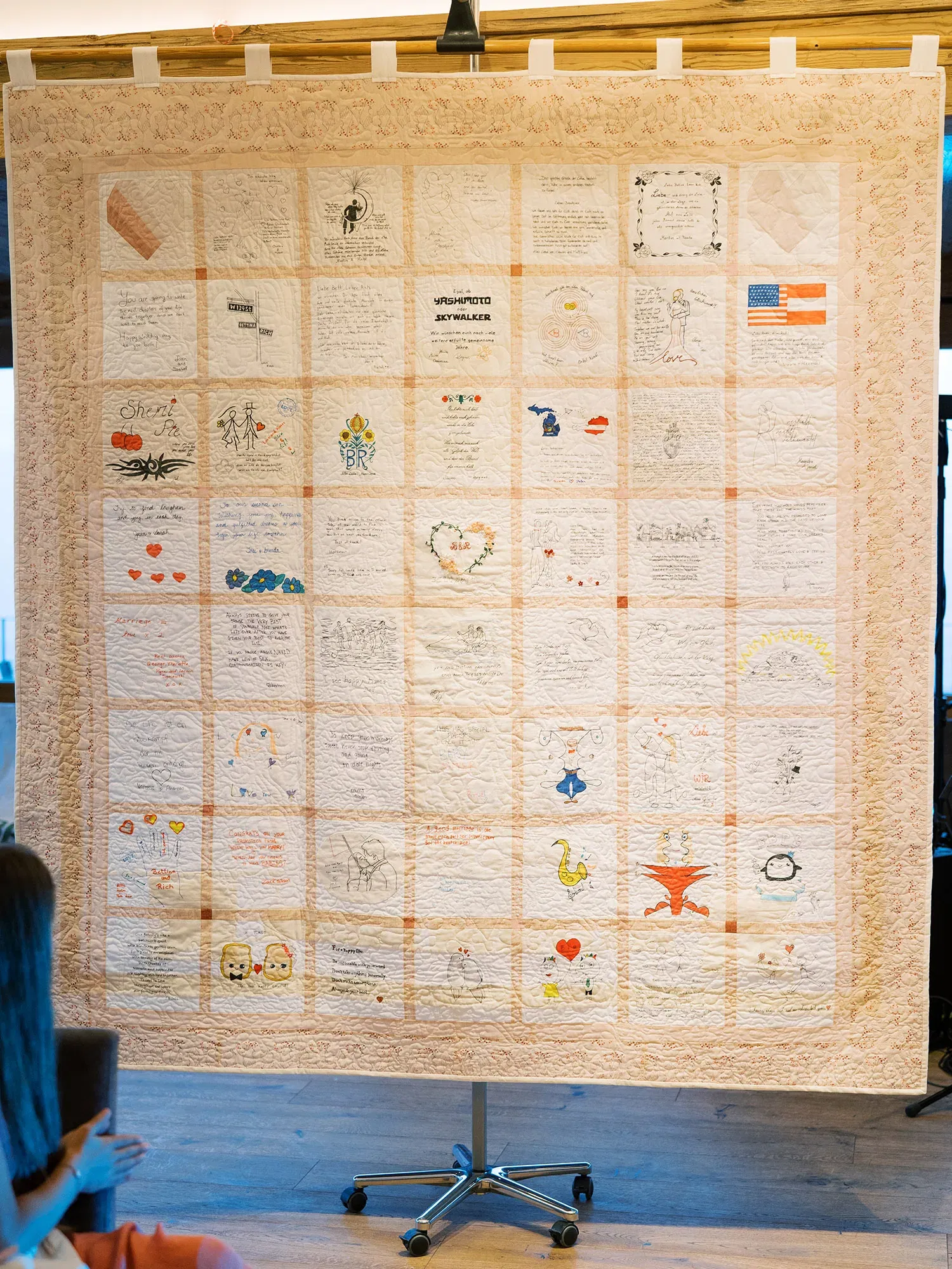 Detailreicher Quilt mit persönlichen Nachrichten und bunten Zeichnungen von Hochzeitsgästen, präsentiert auf einem Ständer als Erinnerung an den besonderen Tag.