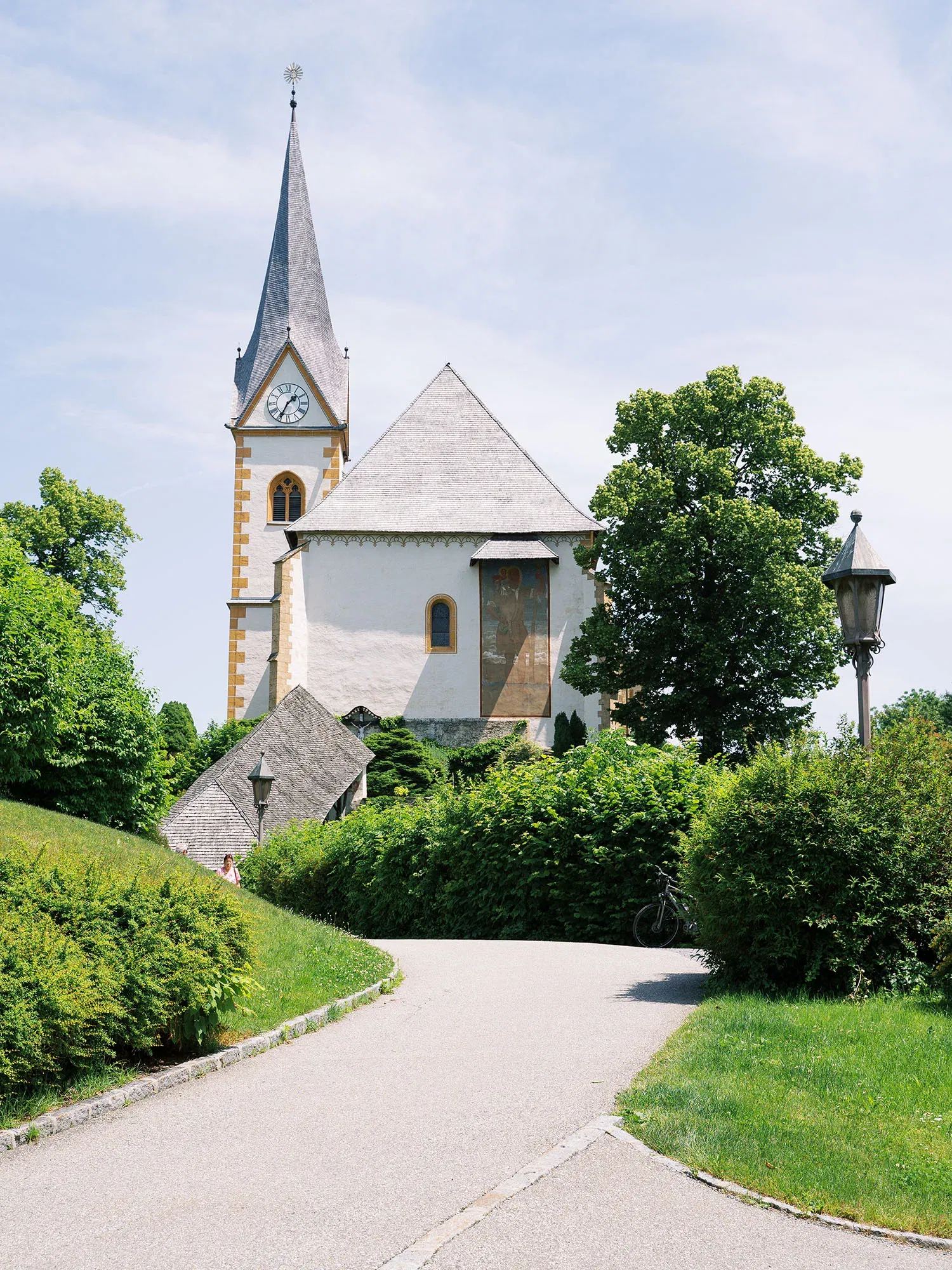 Weiß gestrichene Kirche mit steilem Glockenturm, umgeben von üppigem Grün, mit einem geschwungenen Gehweg, der zur Kirche führt.