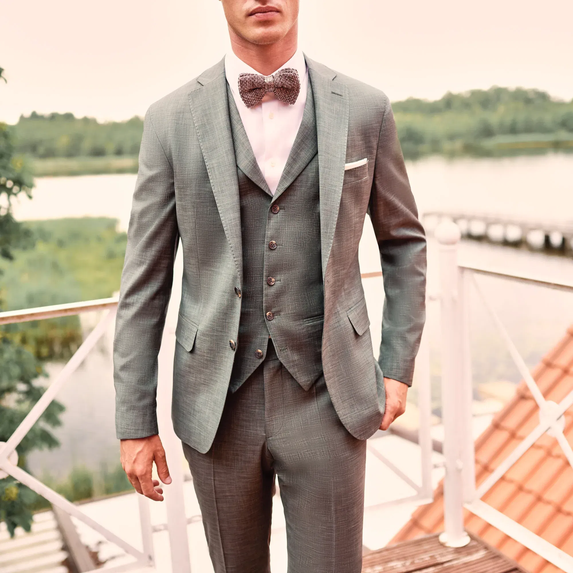 Der Bräutigam trägt einen dreiteiligen grauen Anzug mit einer gemusterten Fliege und steht vor einer idyllischen Naturkulisse auf einer Terrasse am Wasser.