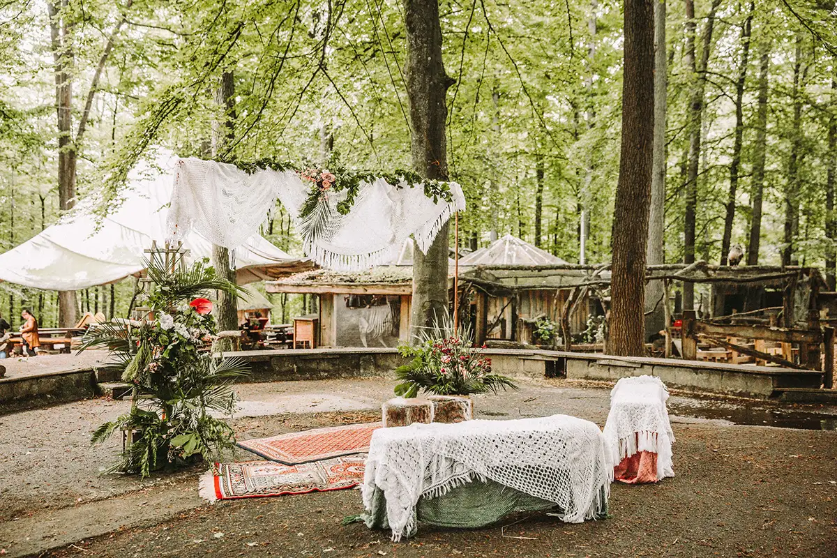 Eine idyllische Freiluft-Hochzeitszeremonie im Wald unter einem mit Blumen geschmückten Baldachin, der rustikale, handgeknüpfte Teppiche und sanfte Naturstimmung betont.