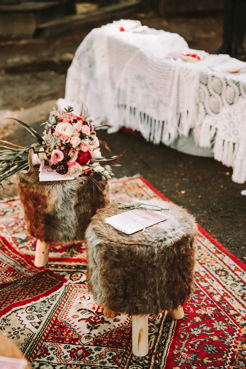 Ein mit Fell überzogener Hocker steht auf einem dekorativen, traditionellen Teppich, darauf ein Blumenstrauß in leuchtenden Farben. Im Hintergrund befindet sich ein mit Spitzenstoff bedeckter Tisch, passend zur Boho-Hochzeit im Wald.