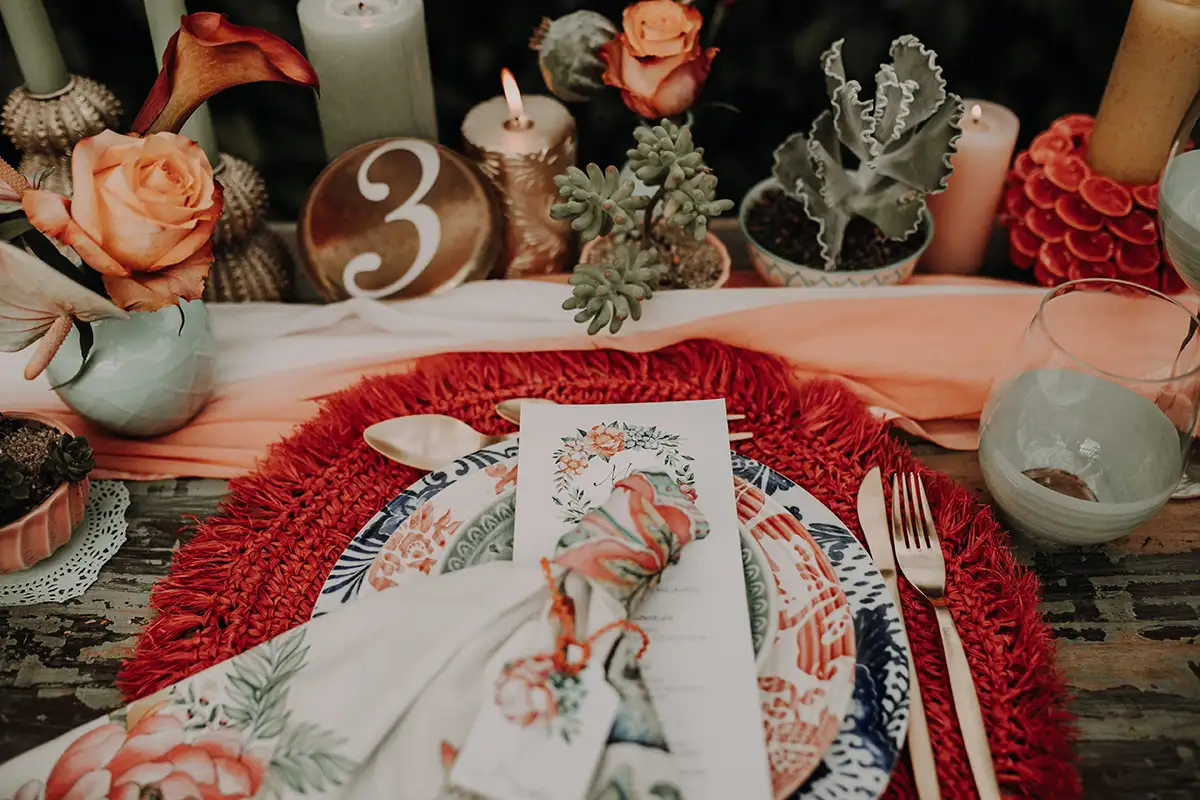 Ein geschmackvoll gedeckter Hochzeitstisch, auf dem farbenfrohes Geschirr, Kerzen und Sukkulenten arrangiert sind, begleitet von orangefarbenen Blumen.