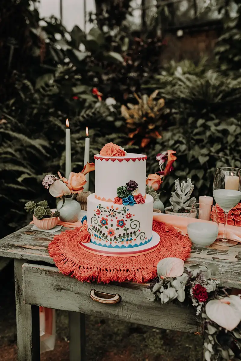 Eine kunstvolle zweistöckige Hochzeitstorte, verziert mit bunten Blumenmotiven, platziert auf einem rustikalen Holztisch und umgeben von Blumendekoration und Kerzen.