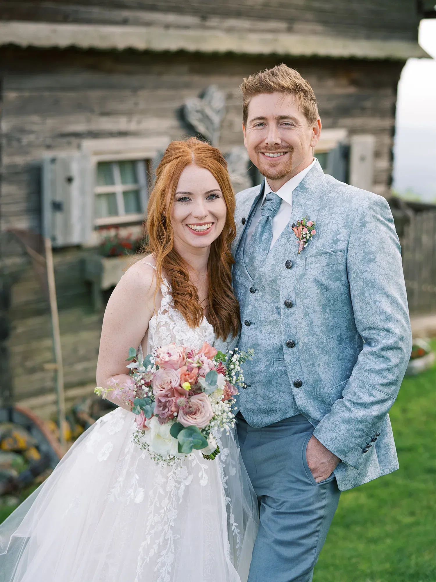 Glückliches Hochzeitspaar vor einem ländlichen, rustikalen Hintergrund, die Braut in einem zarten Brautkleid mit Blumenstrauß, der Bräutigam in einem stilvollen grauen Anzug.